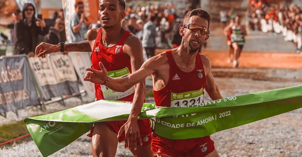 A male runner crossing the finish line at the Mosteiro dos Jerónimos in Belém Lisbon, arms raised in triumph, blue race carpet, crowd cheering, realistic sports photography, daytime