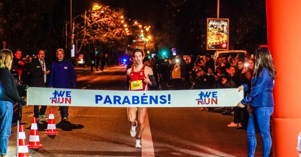 Male runner crossing marathon finish line in Porto park setting, arms raised, chip timer visible, crowd cheering, autumn daylight, realistic sports photography