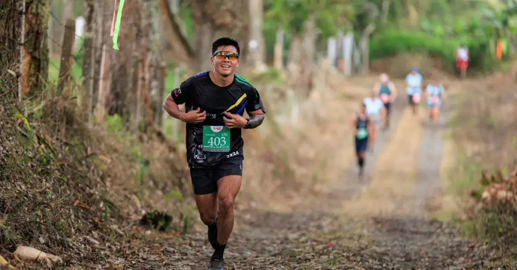 A male trail runner ascending a steep mountain path surrounded by pine forest, wearing a hydration vest and trail shoes, intense effort expression, golden hour lighting, realistic stock photo